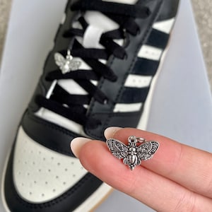 Könnte beinhalten: Ein silberner Motten-Anhänger mit filigranen Details. Der Anhänger hängt an einem schwarzen Schnürsenkel an einem Paar schwarz-weißen Adidas-Schuhen.