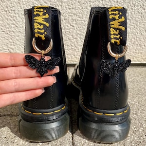 Könnte beinhalten: Schwarze Lederstiefel mit einem gelben und schwarzen Schleifen-Zugband, auf dem "Dr. Martens" steht, und einem schwarzen Schmetterlingsanhänger.