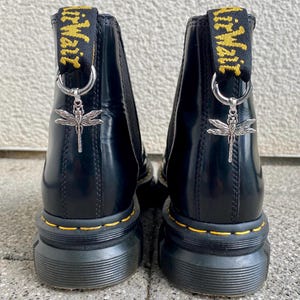 Könnte beinhalten: Schwarze Lederstiefel mit Knöchelriemen, dicker schwarzer Gummisohle und gelben Nähten. Die Stiefel haben einen silbernen Libellenanhänger, der an einem schwarzen Band mit dem Text "Dr. Martens" in Gelb befestigt ist.