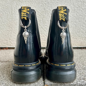 Könnte beinhalten: Schwarze Leder-Ankle Boots mit gelben Nähten und dicker Sohle. Jeder Stiefel hat eine schwarze und gelbe Zuglasche mit der Aufschrift "Air Wair" und einen silbernen Anhänger in Form eines Vogelschädels.
