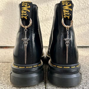 Könnte beinhalten: Paar schwarze Leder-Ankle Boots mit klobiger Sohle. Jeder Stiefel hat einen goldfarbenen Ring und einen silbernen Schwertanhänger, der an der Zuglasche befestigt ist. Die Stiefel haben gelbe Nähte um die Sohle.