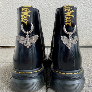 Könnte beinhalten: Schwarze Lederstiefel mit gelben Nähten und einem schwarzen und goldenen Band mit dem Text "Grimmoire" darauf. Jeder Stiefel hat einen silbernen Anhänger in Form eines Nachtfalters mit einem Totenkopf darauf.
