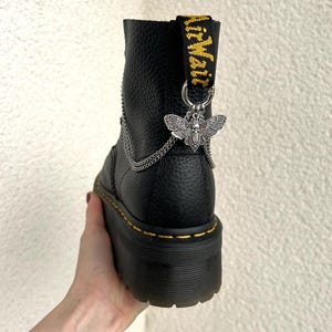 Könnte beinhalten: Schwarzer Leder-Plateau-Stiefel mit silberfarbenem Motten-Charm und Ketten-Detail. Der Stiefel hat eine gelb-schwarze Zuglasche mit dem Markennamen. Die Sohle ist dick und hat gelbe Nähte.