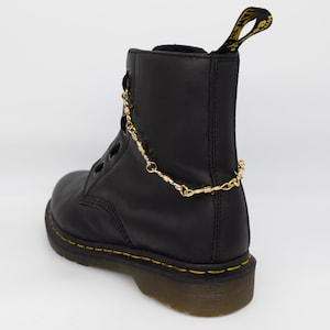 Könnte beinhalten: Schwarze Lederstiefel mit gelben Nähten und einem goldenen Kettendetail. Die Stiefel haben eine dicke Gummisohle.