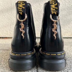 Könnte beinhalten: Paar schwarze Leder-Ankle Boots mit gelben Nähten und dicken Sohlen. Jeder Stiefel hat einen goldfarbenen Anhänger mit einem Caduceus-Symbol, der an der Zugschlaufe hängt.