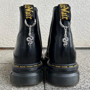 Könnte beinhalten: Schwarze Leder-Ankle Boots mit gelben Nähten und dicken Sohlen. Jeder Stiefel hat eine gelb-schwarze Zuglasche mit der Aufschrift "Air Wair" und einen silbernen Schlangenanhänger.