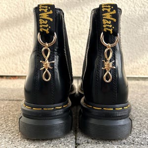 Könnte beinhalten: Schwarze Stiefeletten mit goldfarbenen Stacheldraht-Anhängern. Die Stiefel haben eine dicke, geriffelte Sohle mit gelben Nähten. Der Markenname "AirWair" ist auf der schwarzen Stoffschlaufe zu sehen.