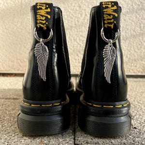 Könnte beinhalten: Schwarze Leder-Ankle Boots mit silbernen Flügel-Anhängern an den Fersenschlaufen. Die Stiefel haben eine klobige Sohle mit gelben Nähten und die Worte "Air Wair" in Gelb auf der Zuglasche.