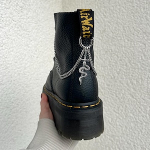 Könnte beinhalten: Schwarzer Leder-Plateau-Stiefel mit gelben Nähten und einem silbernen Ketten- und Schlangenanhänger. Der Stiefel trägt den Markennamen "AirWair" in Gelb auf der Zuglasche.