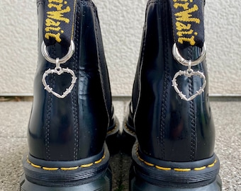 Puede incluir: Botas negras de cuero con detalles de costura amarilla y un dije de corazón de alambre de púas. Las botas tienen una lengüeta de tiro con el texto "Dr. Martens" impreso en amarillo.