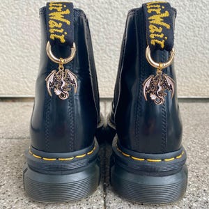 Könnte beinhalten: Schwarze Chelsea-Stiefeletten aus Leder mit gelben Nähten und Zuglaschen. Jeder Stiefel hat einen goldfarbenen Drachenanhänger. Die Stiefel haben dicke, geriffelte Sohlen.