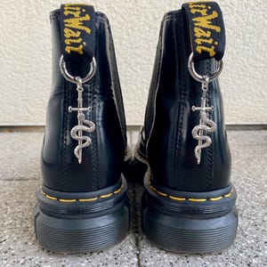Könnte beinhalten: Schwarze Lederstiefel mit dicker Plateau-Sohle und einem gelben und schwarzen Zugband mit der Aufschrift "Dr. Martens". Die Stiefel haben einen silbernen Anhänger mit einer Schlangen- und Schwertmuster.