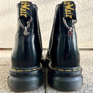 Könnte beinhalten: Paar schwarze Leder-Ankle Boots mit dicker Sohle und gelben Nähten. Jeder Stiefel hat eine schwarz-gelbe Zuglasche mit dem Wort "AirWair" und einem silbernen Herzanhänger.