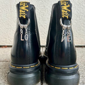 Könnte beinhalten: Schwarze Stiefeletten mit dicker Sohle und gelben Nähten. Die Stiefel haben eine Zuglasche mit gelbem Text und silbernen Anhängern mit den Buchstaben "LO" und "VE".