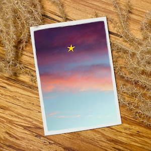 Könnte beinhalten: Ein rechteckiger Druck mit einem farbenfrohen Himmel und einem gelben Stern. Der Himmel wechselt von tiefem Lila zu Orange- und Blautönen. Der Druck liegt auf einer Holzoberfläche, mit getrockneten Pflanzen.
