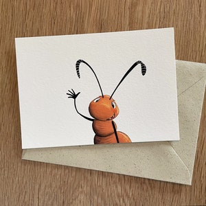 Peut inclure: Carte de vœux avec une fourmi de dessin animé qui salue. La fourmi est orange avec des antennes et des membres noirs, sur fond blanc. Une enveloppe en papier kraft est partiellement visible. La carte et l'enveloppe reposent sur une surface en bois.