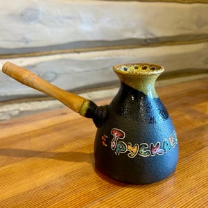 Pode incluir: Uma cafeteira de cerâmica preta com uma pega de madeira. A cafeteira tem um esmalte amarelo e preto e a palavra "русская" escrita em letras coloridas no lado.