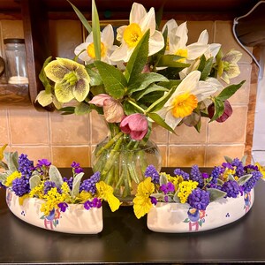Könnte beinhalten: Ein Blumenarrangement mit einer Vase aus klarem Glas mit weißen und gelben Narzissen und zwei cremefarbenen, bootsförmigen Pflanzgefäßen, die mit lila, gelben und grünen Blumen gefüllt sind. Im Hintergrund eine geflieste Wand.