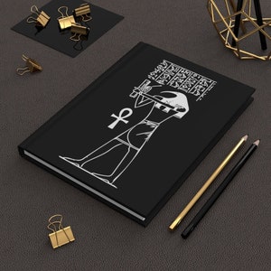 以下が含まれることがあります： エジプトの人物が杖と象形文字を持っている白いイラストが描かれた黒いジャーナル。アンクのシンボルも表示されています。金と黒の鉛筆と金のクリップが暗い表面にあります。