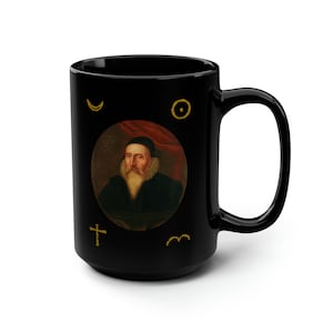 May include: Black ceramic mug with a portrait of a man with a beard and a white collar. The mug has gold symbols on it, including a crescent moon, a circle, a cross, and a squiggly line.