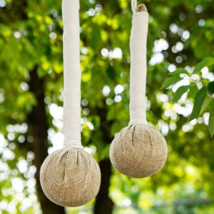 Può includere: Due sfere per massaggi alle erbe realizzate in tessuto testurizzato marrone chiaro, ciascuna fissata a un lungo manico cilindrico avvolto da una spessa corda di cotone bianco sporco. I due oggetti pendono verticalmente con un aspetto intrecciato.