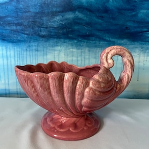 May include: A pink ceramic planter with a seashell design. The planter has a scalloped rim, a pedestal base, and a curved handle. The color is a muted pink with lighter highlights. The background is a blue and white abstract painting.