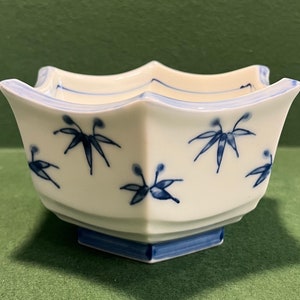May include: A white ceramic bowl with a blue floral design. The bowl has a unique, angular shape with a scalloped rim and a square base. The blue floral pattern is repeated around the bowl's exterior. The bowl is likely for decorative or serving purposes.