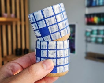 May include: Two small ceramic cups stacked, featuring a blue and white grid pattern. The cups have a speckled, natural-colored base. The design is a modern take on traditional pottery.