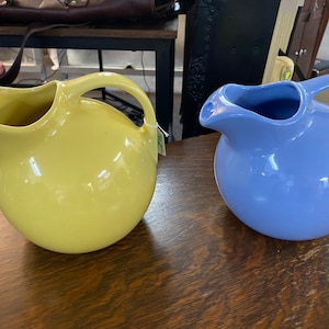 May include: Two ceramic pitchers, one yellow and one periwinkle blue, sit side-by-side on a wooden surface. Each pitcher has a rounded body, a spout, and a curved handle. The pitchers appear to be vintage or antique.