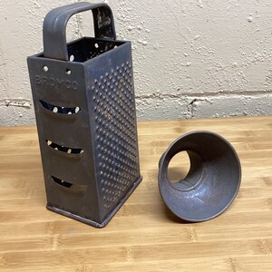 May include: A vintage metal grater with a handle and the word "BROMCO" stamped on the side. The grater is dark gray and has a rectangular shape with multiple grating surfaces. A separate, cone-shaped piece sits beside it.