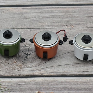 Może przedstawiać: Trzy miniaturowe garnki do gotowania ryżu w różnych kolorach: zielonym, pomarańczowym i białym. Każdy garnek ma srebrną pokrywkę z czarnym przyciskiem na górze.