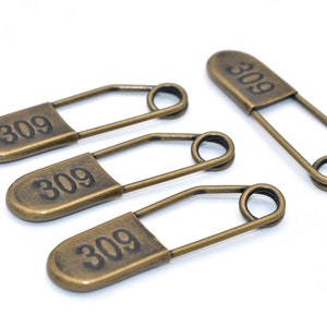 Può includere: Quattro spille di sicurezza in oro antico con il numero 309 stampato su ciascuna. Le spille di sicurezza sono disposte in fila su una superficie bianca.