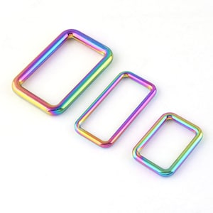 Pode incluir: Três anéis de metal retangulares em tamanhos variados, exibindo um vibrante acabamento iridescente arco-íris. Os anéis estão dispostos diagonalmente contra um fundo branco, mostrando um espectro de cores do amarelo ao azul. Estes anéis são provavelmente usados para artesanato ou joalheria.