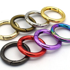 Puede incluir: Una colección de anillos redondos de metal con resorte en varios colores, incluyendo dorado, plateado, bronce, negro, rojo, morado y arcoíris iridiscente. Estos anillos están diseñados con una puerta con resorte para una fácil fijación.