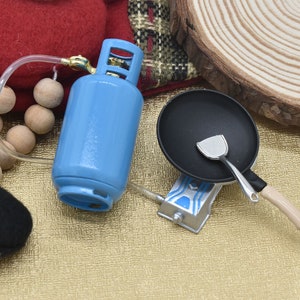 May include: A miniature blue propane tank with a hose and a black frying pan with a spatula on a stovetop.