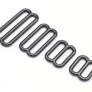 Può includere: Quattro fibbie scorrevoli in metallo nero di diverse dimensioni. Le fibbie sono lisce e hanno una finitura lucida.