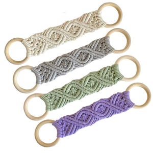 May include: Five macrame wall hangings with wooden rings. The hangings are in various colors, including cream, gray, green, and purple. Each hanging features a diamond pattern.
