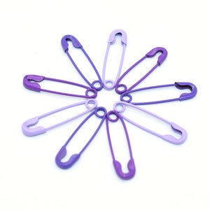 May include: A set of eight safety pins arranged in a starburst pattern. The pins are all different shades of purple, ranging from light lavender to deep violet.