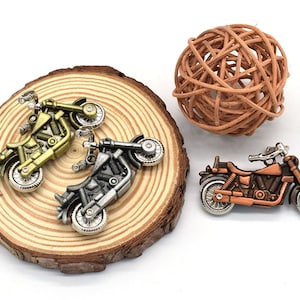 Könnte beinhalten: Drei Miniatur-Motorrad-Anhänger in Gold-, Silber- und Kupfer-Finish. Die Anhänger werden auf einer Holzoberfläche mit einem geflochtenen Ball im Hintergrund präsentiert.
