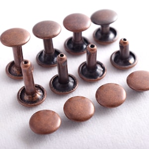 Op de afbeelding: Een verzameling van 11 koperkleurige metalen klinknagels, sommige met een verhoogde kop en andere met een platte kop. De klinknagels zijn gerangschikt op een wit oppervlak.