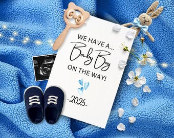 May include: A white card with the text "We have a... Baby Boy on the way! -2025" with blue watercolor hearts. The card is surrounded by a blue blanket, a wooden rattle, a knitted bunny, and white flowers.