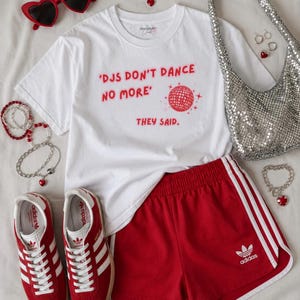 Può includere: Un'elegante composizione con una maglietta bianca con testo rosso e grafica a palla da discoteca, pantaloncini rossi con strisce bianche e scarpe da ginnastica rosse. Accessori: occhiali da sole a forma di cuore, gioielli e borsa argentata con paillettes.