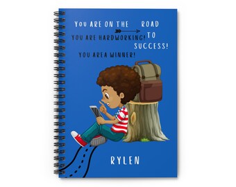 May include: A blue spiral notebook with a cartoon illustration of a young boy sitting on a tree stump with a backpack. The text on the notebook reads "You are on the road to success! You are hardworking! You are a winner!" and "RYLEN".