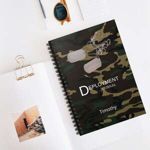 May include: A green, brown, and black camouflage notebook with a spiral binding. The notebook has two silver dog tags hanging from a chain. The text "DEPLOYMENT JOURNAL" and "Timothy" is printed on the front cover.