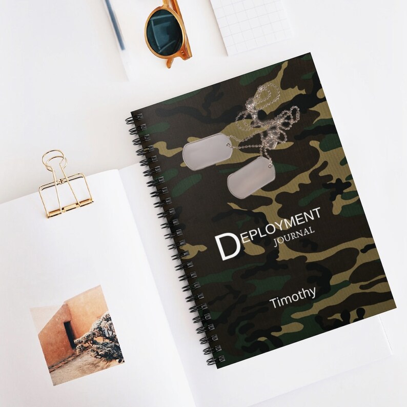 May include: A green, brown, and black camouflage notebook with a spiral binding. The notebook has two silver dog tags hanging from a chain. The text "DEPLOYMENT JOURNAL" and "Timothy" is printed on the front cover.