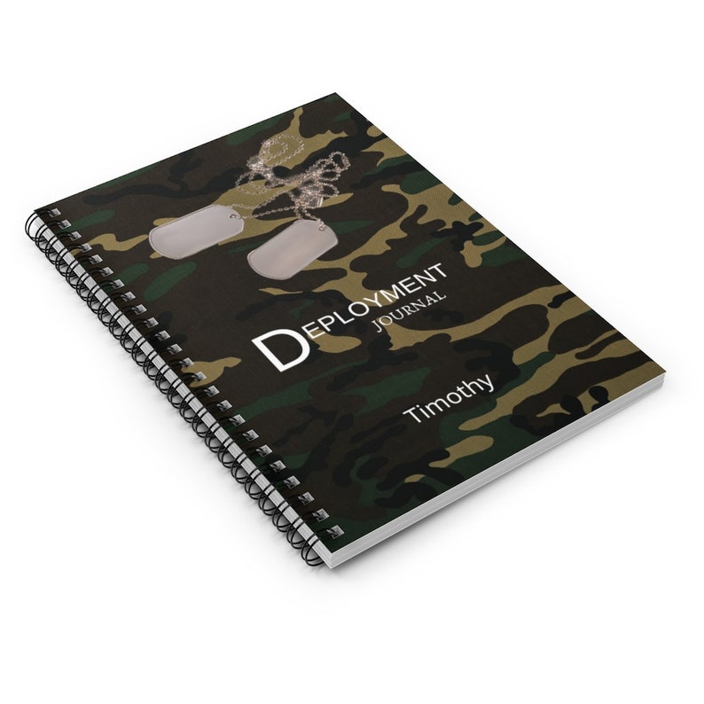 May include: A green, brown, and black camouflage spiral bound notebook with the text "Deployment Journal" and the name "Timothy" printed on the cover. Two silver dog tags are attached to the notebook with a silver chain.