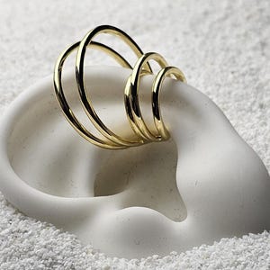 Könnte beinhalten: Ein goldener Ohrklemmen-Schmuck mit drei verbundenen Ringen liegt auf einer weißen ohrförmigen Skulptur, vor einem Hintergrund aus weißem Sand. Das Schmuckstück ist ein modernes Accessoire.