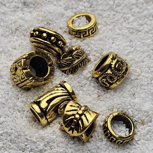 Könnte beinhalten: Eine Sammlung goldfarbener Metallperlen mit verschiedenen Designs, darunter geometrische Muster, florale Motive und geprägte Details. Die Perlen sind auf einer weißen, strukturierten Oberfläche verstreut.