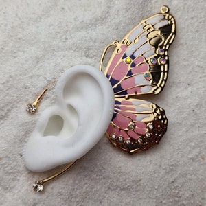 Op de afbeelding: Een gouden vlinder oorring met roze en witte emaille en strass. De oorring is bevestigd aan een gouden oormanchet met een kleine strass.