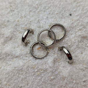 Peut inclure: Cinq boucles d'oreilles créoles argentées disposées sur une surface blanche texturée. Les boucles d'oreilles sont petites, circulaires et présentent une finition polie. L'image est une prise de vue rapprochée, mettant en valeur les détails.